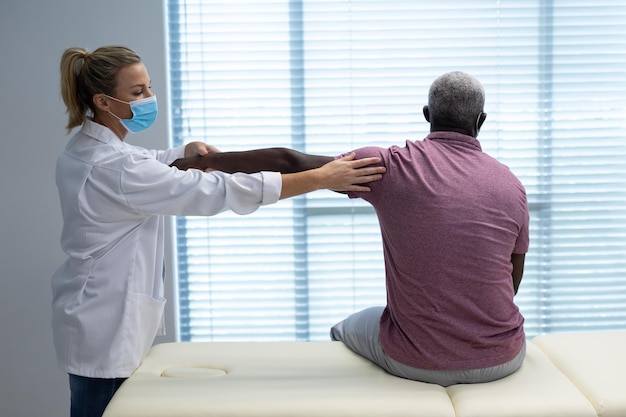 Foto fisioterapeuta caucasiana usando máscara esticando o braço de um paciente afro-americano