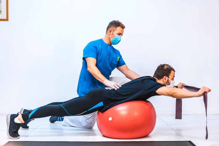 Foto fisioterapeuta com máscara e um paciente fazendo exercícios com uma borracha em uma bola gigante. fisioterapia com medidas de proteção para a pandemia de coronavírus, covid-19. osteopatia, massagem esportiva