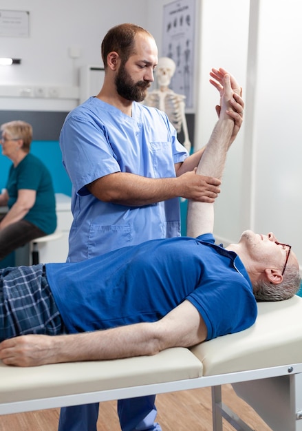 Fisioterapeuta dando masaje quiropráctico a un anciano, ayudando con la recuperación de los músculos del brazo. Asistente de masaje para estirar la mano de la persona mayor para el tratamiento y cuidado de la cinetoterapia.