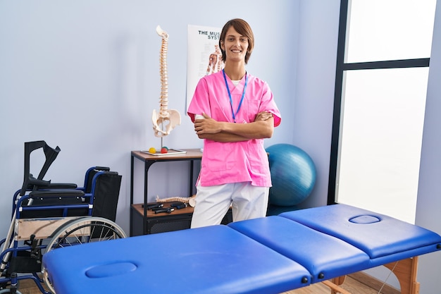 Fisioterapeuta jovem e bonita mulher hispânica sorrindo confiante em pé com os braços cruzados gesto na clínica de reabilitação