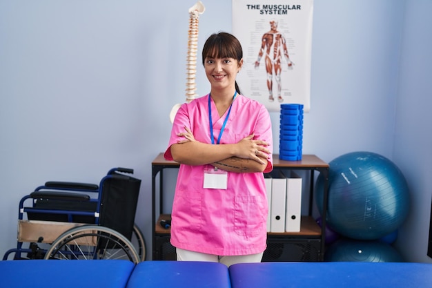 Fisioterapeuta jovem e bonita mulher hispânica sorrindo confiante em pé com os braços cruzados gesto na clínica de reabilitação