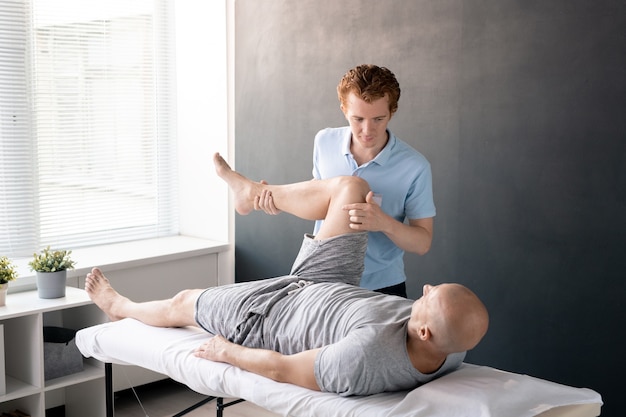 Fisioterapeuta sério olhando para seu paciente com a perna dobrada no joelho enquanto o ajuda com um dos exercícios físicos no centro de reabilitação