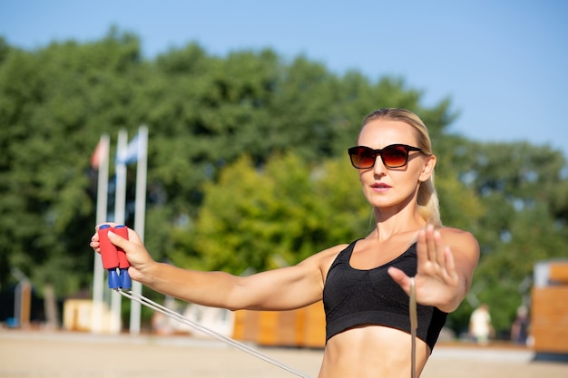 Fit blondes Mädchen mit Sonnenbrille beim Stretching mit Springseil am Strand