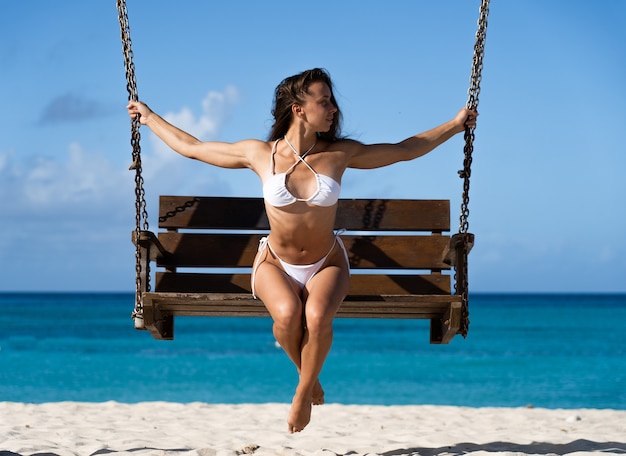 Fit Frau im weißen Bikini, der am Strand sitzt und Sonnenbad weißen Sand und klares azurblaues Meerwasserkonzept von sorglosem Urlaub und Freizeit genießt