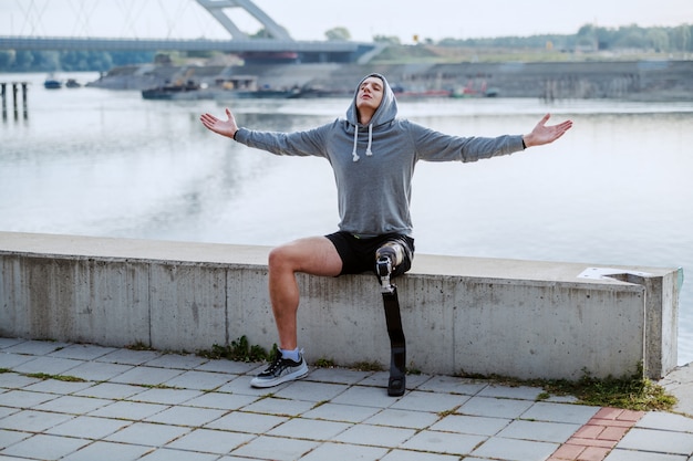 Fit gesunden kaukasischen behinderten Sportler mit künstlichem Bein und Kapuzenpulli auf dem Kopf sitzen auf Kai mit geöffneten Armen.