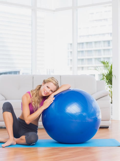 Fit loira sentada ao lado de bola de exercício sorrindo para a câmera