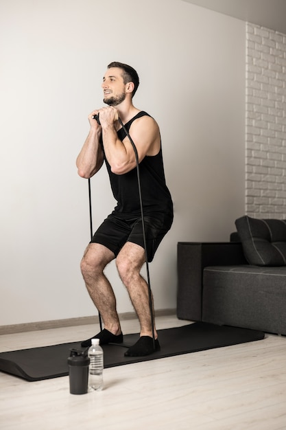 Fit man führt Übungen mit einem Widerstandsband durch. Hartes Training zu Hause. Minimalistisches Interieur im Hintergrund. Wasserflasche in der Nähe von Yogamatte. Ein Mann hat Freude daran, seine Lieblingsübung zu machen.