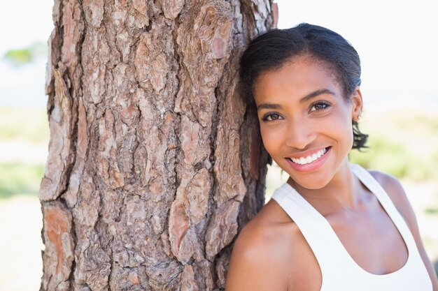 Fit mujer apoyada contra árbol sonriendo a la cámara