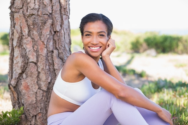 Fit mujer sentada contra el árbol sonriendo a la cámara