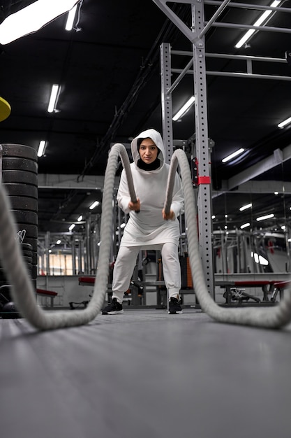 Fit muslimische Frau im sportlichen Hijab, der Crossfit-Übungen mit Seil im modernen Fitnessstudio macht, arabisches Frauentraining allein, im Sport engagiert