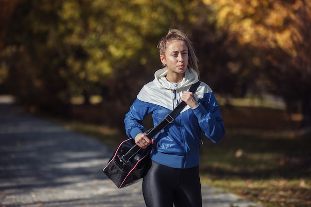 Fit schlanke Frau in Sportbekleidung und eine Trainingstasche auf der Schulter geht beim Training durch den Herbstpark