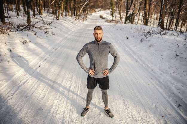 Fit Sportler, der in der Natur mit den Händen auf den Hüften am verschneiten Wintertag steht.