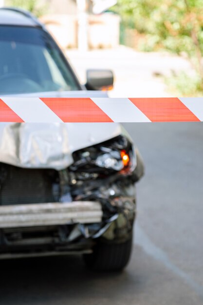 Fita vermelha de advertência cruzada na frente do acidente de carro. Capô quebrado do carro na estrada cercada por fita vermelha de advertência. Pára-choque quebrado e faróis de carro com luz no asfalto da superfície da estrada com ninguém.