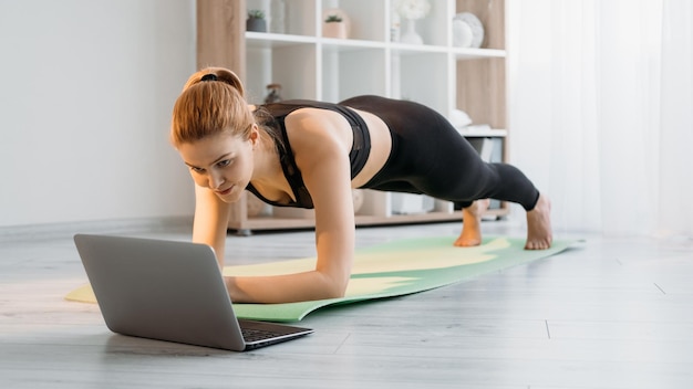 Fitness en casa Pilates en línea Sport video tutorial Mujer atlética en ropa deportiva sosteniendo tablón haciendo ejercicio físico con computadora portátil en colchoneta de yoga en el piso en el interior de la sala de luz