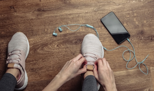 Fitness, concepto de deporte. Mujer atarse los cordones de los zapatos deportivos para correr mientras está sentado en el suelo con un teléfono inteligente y auriculares.