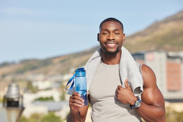 Fitness de retrato e garrafa de água com um homem negro esportivo ou corredor em pé ao ar livre com uma toalha durante o exercício Treinamento de corrida e treino com um jovem atleta do sexo masculino do lado de fora para cardio