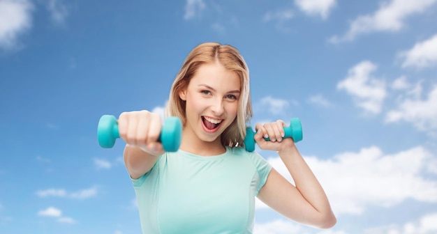 fitness, deporte, ejercicio y concepto de la gente - hermosa mujer deportiva sonriente con mancuerna en pie de lucha sobre el cielo azul y el fondo de las nubes