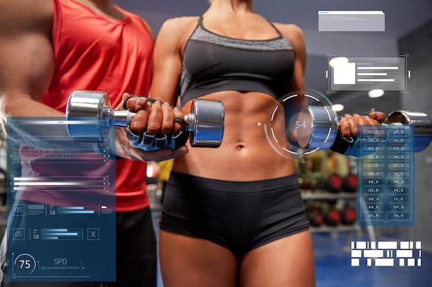 Foto fitness, deporte, ejercicio, personas y concepto de levantamiento de pesas: mujer joven y entrenador personal con pesas flexionando músculos en el gimnasio sobre gráficos virtuales