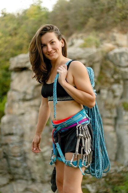 Fitness, deporte extremo y concepto de estilo de vida saludable: el escalador sostiene la cuerda. Chica delgada tiene equipo de escalada en una pared de roca. y mirando a cámara sobre fondo de roca