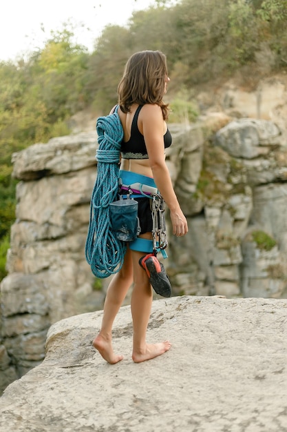 Fitness, deporte extremo y concepto de estilo de vida saludable: el escalador sostiene la cuerda. Chica delgada tiene equipo de escalada en una pared de roca. y mirando a cámara sobre fondo de roca