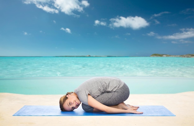 fitness, deporte, gente y concepto de estilo de vida saludable - mujer feliz haciendo yoga en pose de niño en la alfombra sobre el fondo del mar y el cielo