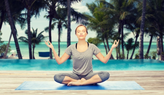 fitness, deporte, gente y concepto de estilo de vida saludable - mujer haciendo meditación de yoga en posición de loto en la alfombra sobre la piscina del hotel en el fondo de la playa tropical