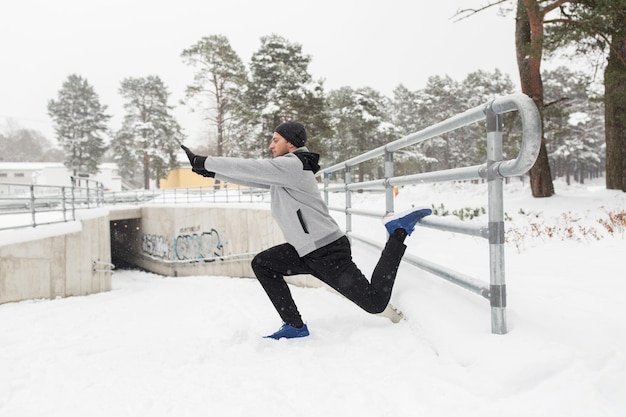 fitness, deporte, gente, ejercicio y concepto de estilo de vida saludable - joven estirando la pierna y calentándose en la cerca en invierno