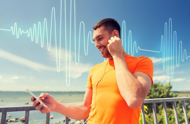 fitness, deporte, gente, tecnología y concepto de estilo de vida saludable - joven sonriente con smartphone y auriculares escuchando música en la playa de verano sobre ondas de sonido o diagrama de señal