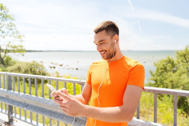 fitness, deporte, gente, tecnología y concepto de estilo de vida saludable - joven sonriente con smartphone y auriculares escuchando música en la playa de verano