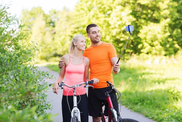 fitness, deporte, gente, tecnología y concepto de estilo de vida saludable - pareja feliz con bicicleta tomando fotos por teléfono inteligente en selfie stick al aire libre