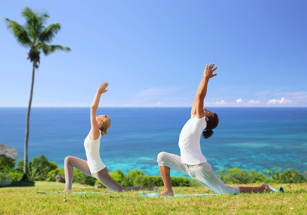 fitness, deporte, relajación y concepto de personas: pareja haciendo yoga en pose de estocada baja sobre un fondo natural exótico con palmeras y océano