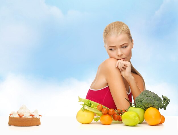 fitness, dieta, salud y concepto de comida - mujer que duda con frutas y pastel