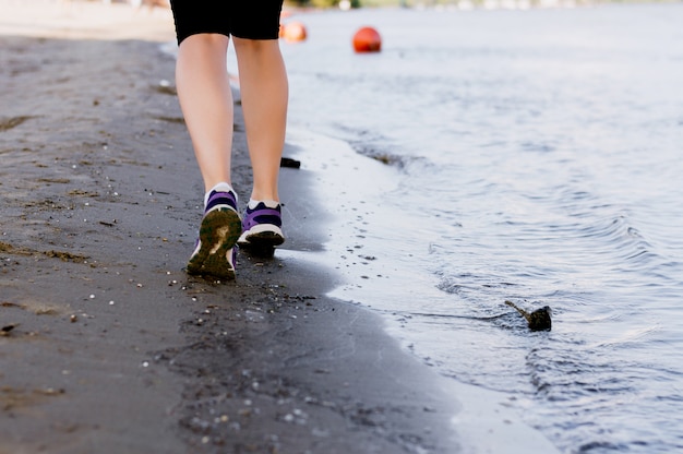 Fitness e correr na praia, pernas de corredor de mulher em sapatos na areia perto do mar