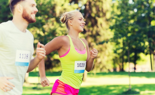 Foto fitness, esporte, amizade, corrida e conceito de estilo de vida saudável - amigos adolescentes felizes ou casal de desportistas correndo maratona com números de distintivo ao ar livre