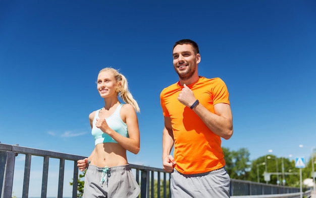 fitness, esporte, amizade e conceito de estilo de vida saudável - casal sorridente com relógio de freqüência cardíaca correndo à beira-mar de verão