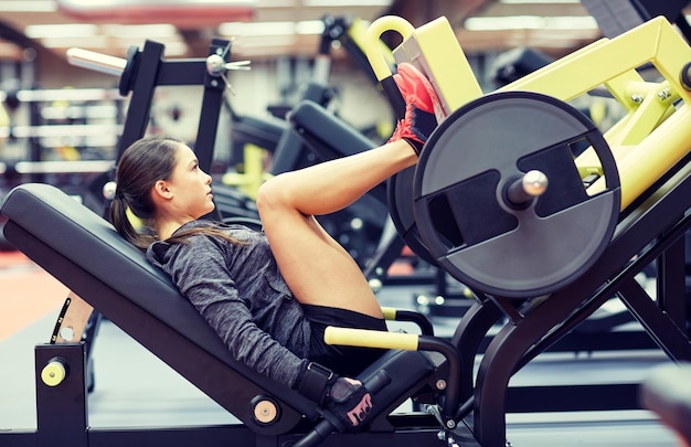 fitness, esporte, musculação, exercício e conceito de pessoas - jovem mulher flexionando os músculos na máquina de prensa de pernas no ginásio