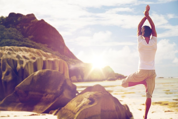 fitness, esporte, pessoas e conceito de estilo de vida saudável - homem meditando em pose de árvore de ioga sobre fundo de praia exótica de costas