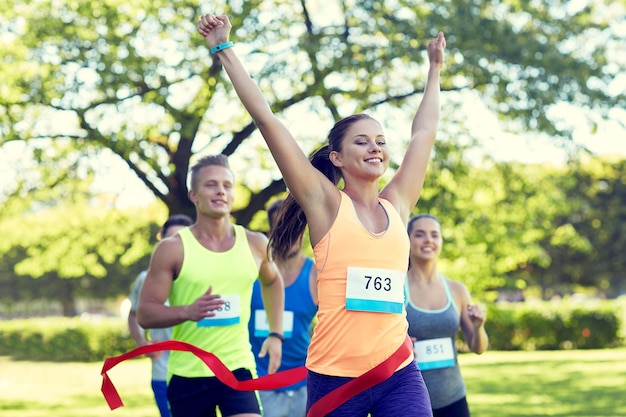 fitness, esporte, vitória, sucesso e conceito de estilo de vida saudável - mulher feliz vencendo corrida e chegando primeiro a terminar a fita vermelha sobre grupo de desportistas correndo maratona com números de distintivo ao ar livre