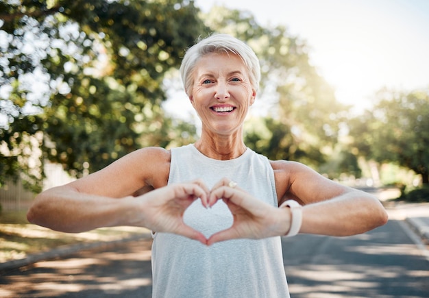 Foto fitness feliz e mãos de coração de velha na natureza depois de correr para saúde, bem-estar e treino sorrir motivação e paz com senhora sênior e sinal de amor, fé e treinamento na natureza