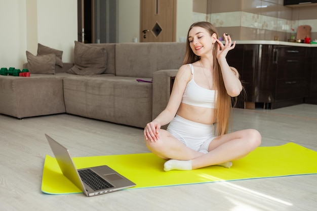 Fitness-Frau in Sportkleidung schaut in den Laptop-Bildschirm und sieht sich ein Fitness-Tutorial an, während sie auf der Matte im Wohnzimmer sitzt Sport zu Hause