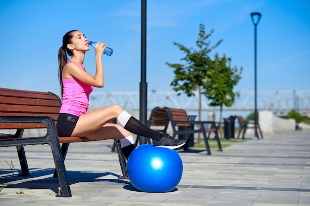 Fitness-Frau mit Flasche und Gummi-Fit-Ball Weibliches Trinkwasser nach dem Training auf der Bank im Sommerpark