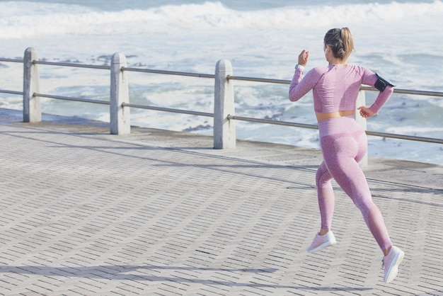 Fitness für Frauen und Laufen auf dem Seeweg im Freien und schnell für Wellness, Gesundheit und Ziele im Sommer Läufermädchen Geschwindigkeit und Gleichgewicht bei Sonnenschein mit tragbarer Smartphone-Technologie und Wellen am Meer in Kapstadt