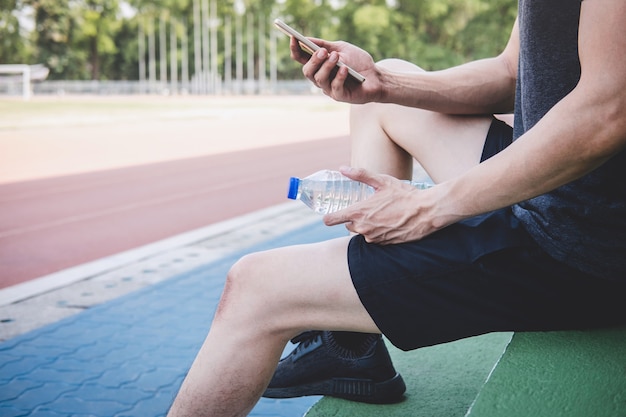 Fitness joven atleta hombre descansando en un banco con una botella de agua preparándose para correr en la pista de carretera
