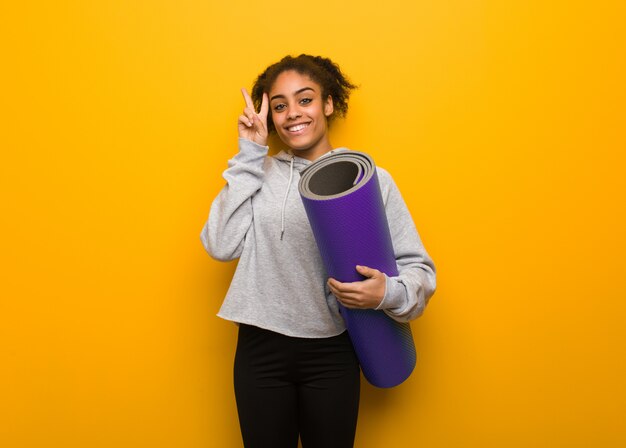 Fitness joven mujer negra divertida y feliz haciendo un gesto de victoria. Sosteniendo una estera.