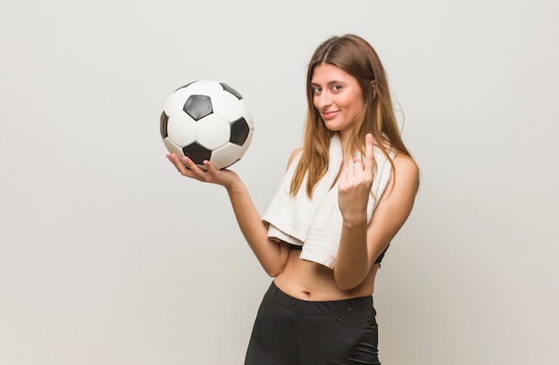 Fitness joven mujer rusa invitando a venir. Sosteniendo un balón de fútbol.