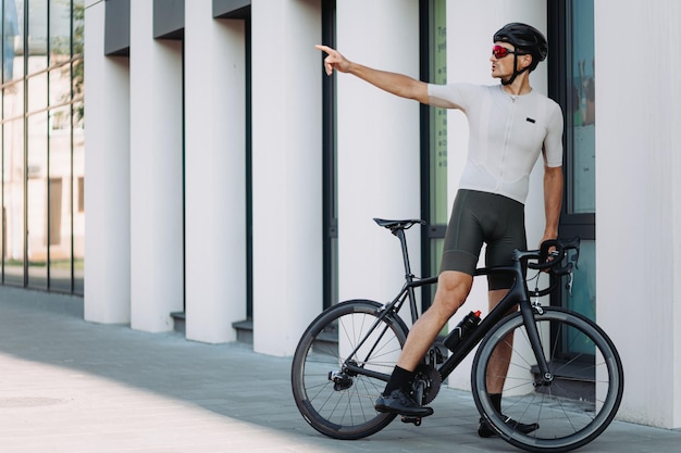 Fitness junger Mann mit Schutzhelm und Brille, der auf einem schwarzen Fahrrad sitzt und mit der Hand zur Seite zeigt Outdoor-Training des kaukasischen Radfahrers Stadtgebiet