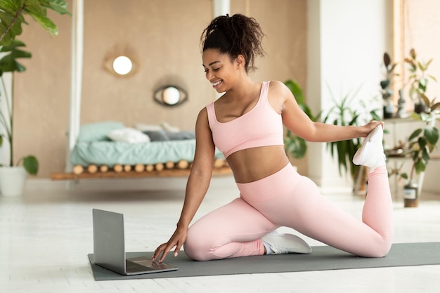 Fitness en línea Mujer negra en forma estirándose en casa sentada en una alfombra de yoga frente a la computadora portátil y sonriendo espacio de copia