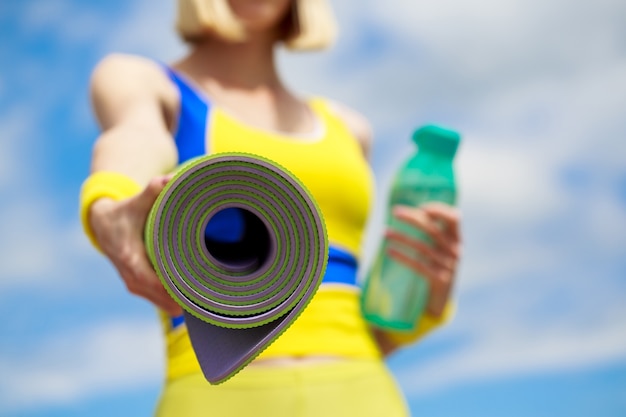 Fitness-Mädchen mit Yogamatte über Himmelhintergrund. Frau in Sportkleidung hält eine Yogamatte und eine Flasche Wasser.