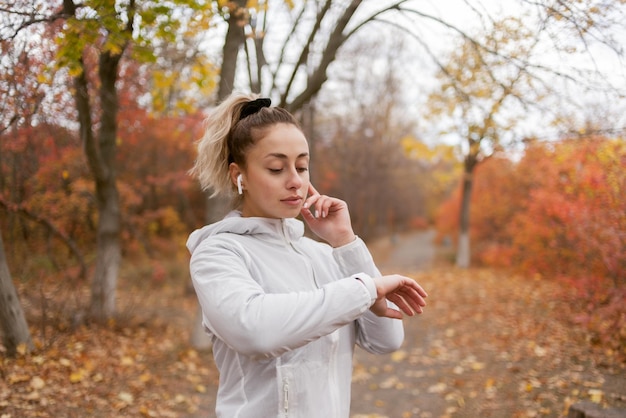 Fitness mujer escucha música con auriculares y mira un reloj inteligente en el bosque de otoño mientras trota Sports Gadgets