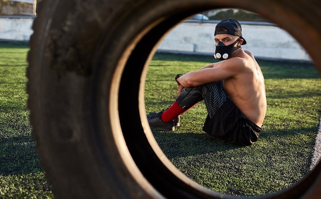 Fitness muskulöser Mann in Maske, um die Belastung der Atemmuskulatur zu erhöhen, die nach Übungen auf Reifen im Freien sitzt Platz für Ihren Text kopieren Hemdloser Sportler, der im Stadion trainiert